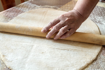 Senior woman hands are rolling out dough in flour with rolling pin in her home kitchen. Homemade noodle or pasta production by grandma.