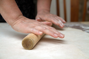 Senior woman hands are rolling out dough in flour with rolling pin in her home kitchen. Homemade noodle or pasta production by grandma.