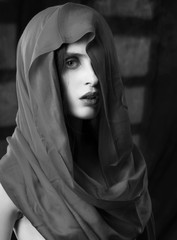 Beautiful girl with fashionable makeup, wearing a silk scarf thrown over her head that covers her naked breast, sensually poses on the background of the shadow from the window. Black and white