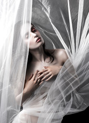 A beautiful slim topless girl poses among the white fabric of a veil that flutters in the wind and that covers her nakedness. Artistic, conceptual, blurry photo.