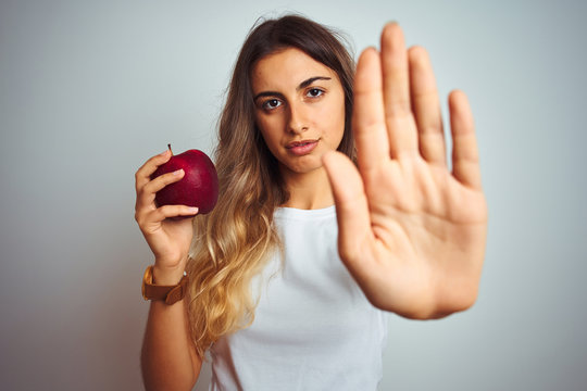 Young beautiful woman eating red apple over grey isolated background with open hand doing stop sign with serious and confident expression, defense gesture