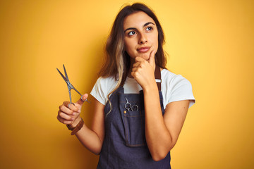 Young beautiful hairdresser woman holding scissors over yellow isolated background serious face...