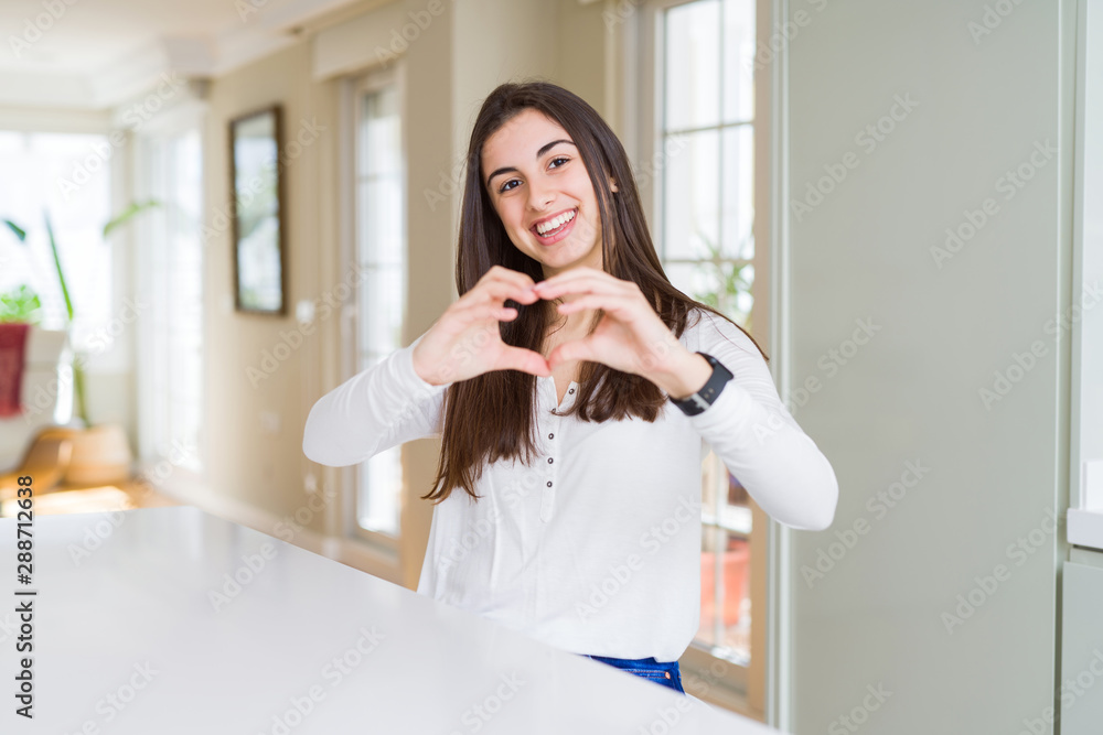 Canvas Prints Beautiful young woman sitting on white table at home smiling in love showing heart symbol and shape with hands. Romantic concept.