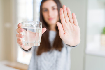 Beautiful young woman drinking a fresh glass of water with open hand doing stop sign with serious...