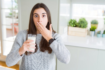 Beautiful young woman drinking a glass of fresh milk cover mouth with hand shocked with shame for...