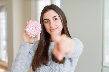 Beautiful young woman eating sugar marshmallow pink donut pointing with finger to the camera and to you, hand sign, positive and confident gesture from the front