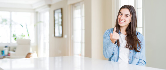 Wide angle picture of beautiful young woman sitting on white table at home Beckoning come here gesture with hand inviting happy and smiling