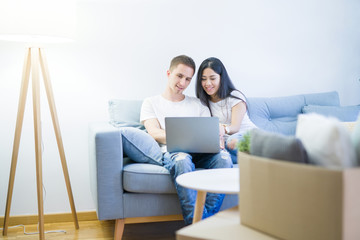 Young beautiful couple sitting on the sofa using laptop at new home around cardboard boxes