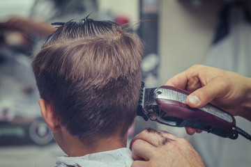 Barber is using clipper to make a styling to Caucasian boy.