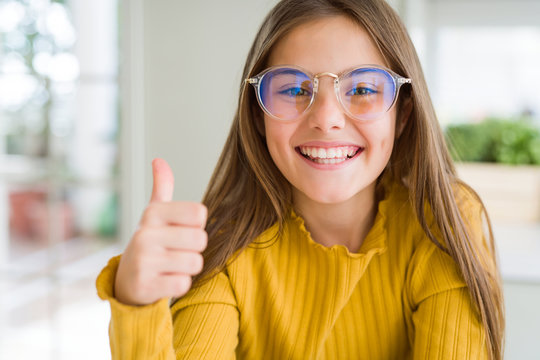 Beautiful young girl kid wearing glasses doing happy thumbs up gesture with hand. Approving expression looking at the camera showing success.