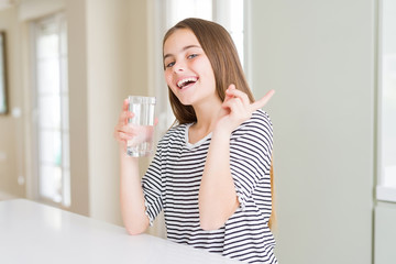 Beautiful young girl kid drinking a fresh glass of water very happy pointing with hand and finger to the side