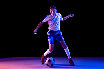 Young boy as a soccer or football player in sportwear making a feint or a kick with the ball for a goal on dark studio background. Fit playing boy in action, movement, motion at game. Purple neon