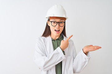 Young chinese engineer woman wearing coat helmet glasses over isolated white background amazed and smiling to the camera while presenting with hand and pointing with finger.