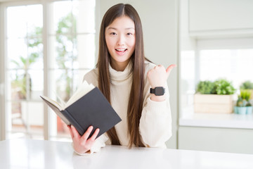 Beautiful Asian woman reading a book pointing and showing with thumb up to the side with happy face smiling