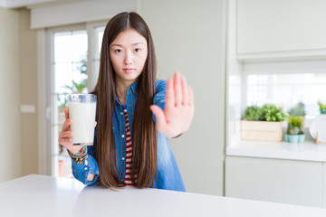 Beautiful Asian woman drinking a fresh glass of milk with open hand doing stop sign with serious and confident expression, defense gesture