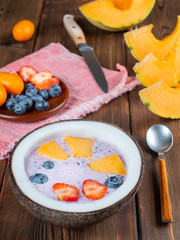 Vertical view of blueberry smoothie in a coconut bowl with strawberries and melon and kumquat on a red towel with a teaspoon and knife. Soft focus.