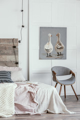 Real photo of a ducks painting hanging on white wall above a gray chair. Bright bedroom interior with pastel bed