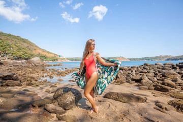 Sexy young woman with long hair in red swimwear with colored beachwear standing on the beach with rocks. Sea vacation. Summer vacation fun. Tropical beach lifestyle. Holiday leisure. Island lifestyle.
