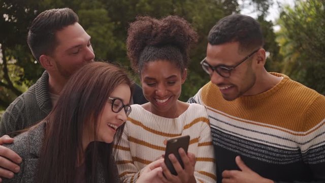 Happy diverse group of friends looking at cellphone enjoying social media phone app technology in park