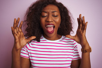 Young african american woman wearing striped t-shirt standing over isolated pink background celebrating mad and crazy for success with arms raised and closed eyes screaming excited. Winner concept
