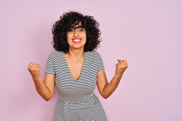 Young arab woman with curly hair wearing striped dress over isolated pink background celebrating surprised and amazed for success with arms raised and open eyes. Winner concept.