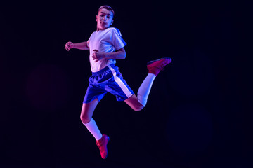 Fototapeta premium Young boy as a soccer or football player in sportwear making a feint or a kick with the ball for a goal on dark studio background. Fit playing boy in action, movement, motion at game. Purple neon