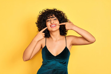 Young arab woman with curly hair wearing elegant dress over isolated yellow background smiling cheerful showing and pointing with fingers teeth and mouth. Dental health concept.