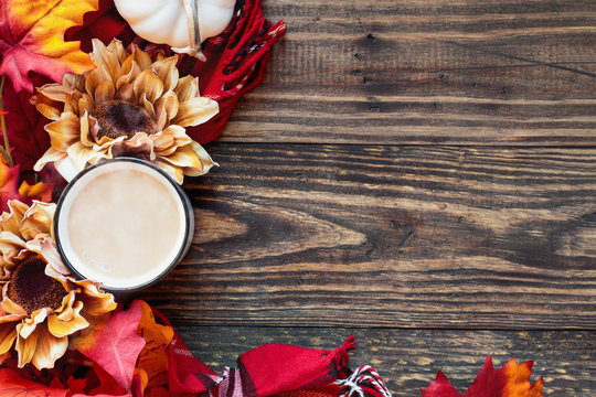 Delicious hot, steaming cup or mug of coffee with cream over a rustic wood table background and surrounded by autumn leaves, red plaid scarf, flowers and white mini pumpkins. 