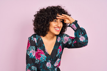 Young arab woman with curly hair wearing floral dress over isolated pink background very happy and smiling looking far away with hand over head. Searching concept. © Krakenimages.com