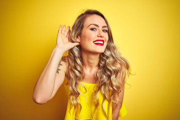 Young attactive woman wearing t-shirt standing over yellow isolated background smiling with hand over ear listening an hearing to rumor or gossip. Deafness concept.