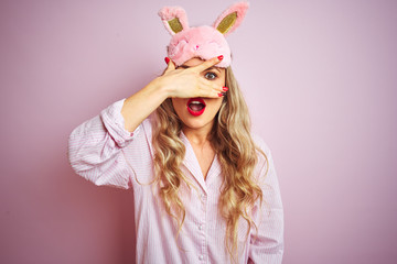 Young beautiful woman wearing pajama and sleep mask over pink isolated background peeking in shock covering face and eyes with hand, looking through fingers with embarrassed expression.