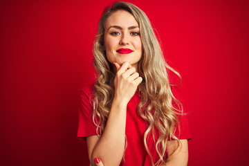 Young beautiful woman wearing basic t-shirt standing over red isolated background looking confident at the camera with smile with crossed arms and hand raised on chin. Thinking positive.