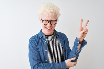 Young albino blond man wearing denim shirt and glasses over isolated white background smiling with happy face winking at the camera doing victory sign with fingers. Number two.