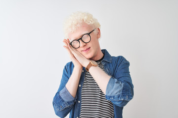 Young albino blond man wearing denim shirt and glasses over isolated white background sleeping tired dreaming and posing with hands together while smiling with closed eyes.