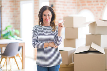 Middle age woman moving to a new house arround cardboard boxes with a big smile on face, pointing with hand and finger to the side looking at the camera.