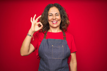 Middle age senior woman wearing apron uniform over red isolated background smiling positive doing...