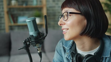 Cheerful young lady in glasses is talking recording audio using microphone in apartment enjoying communication and blogging activity. Youth culture and devices concept.