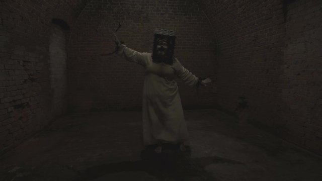 A funny ghost of a fat woman with a rusty cage on his head and with shackles on his hands dances in the gloomy room of old dungeon.