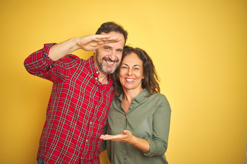 Beautiful middle age couple over isolated yellow background gesturing with hands showing big and large size sign, measure symbol. Smiling looking at the camera. Measuring concept.