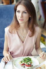 Portret of attractive young woman in a pink dress has lunch in a restaurant. A dish with salad and drinks on the table.