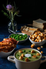 Wooden table with bowls of thick green pea soup embellished with roasted rusks, green peas, slices of grilled bacon, crushed black pepper and cream.