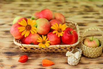 Red ripe apples and autumn flowers lie in a wicker plate on a wooden table. Wicker bast shoes and...