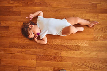 Beautiful toddler child girl wearing white bodysuit lying down on the floor using pacifier