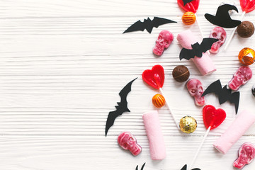 Halloween candy border with skulls, black bats, ghost, spider paper decorations on white wooden background, flat lay. Halloween sweets mockup. Copy space. Trick or treat