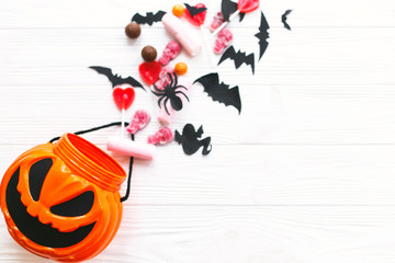 Trick or treat. Halloween candy spilled from jack o lantern bucket with skulls, black bats, ghost, spider decorations on white wooden background, flat lay. Halloween sweets. Copy space.