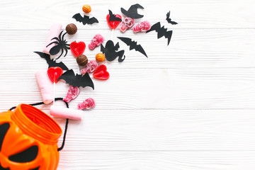 Halloween candy spilled from jack o lantern bucket with skulls, black bats, ghost, spider decorations on white wooden background, flat lay. Halloween sweets. Copy space. Trick or treat