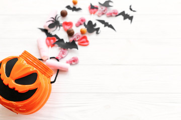 Happy Halloween. Halloween candy spilled from jack o lantern bucket with skulls, black bats, ghost, spider decorations on white wooden background, flat lay. Copy space. Trick or treat