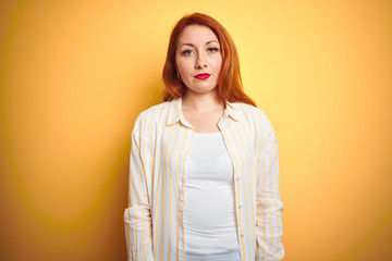 Obraz premium Beautiful redhead woman wearing striped shirt standing over isolated yellow background with serious expression on face. Simple and natural looking at the camera.