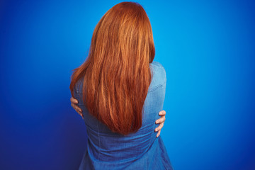 Young beautiful redhead woman wearing denim shirt standing over blue isolated background Hugging oneself happy and positive from backwards. Self love and self care - obrazy, fototapety, plakaty