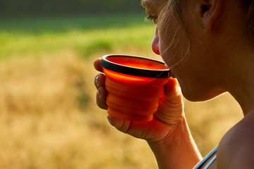 Young woman is enjoying cup of coffee outside with beautiful view of mountains and woods during a sunset/sunrise